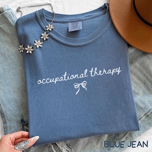 May include: A blue Comfort Colors t-shirt with the words "occupational therapy" in white script and a bow graphic. A floral necklace and a brown hat are also visible. The shirt is folded on top of a pair of blue jeans.