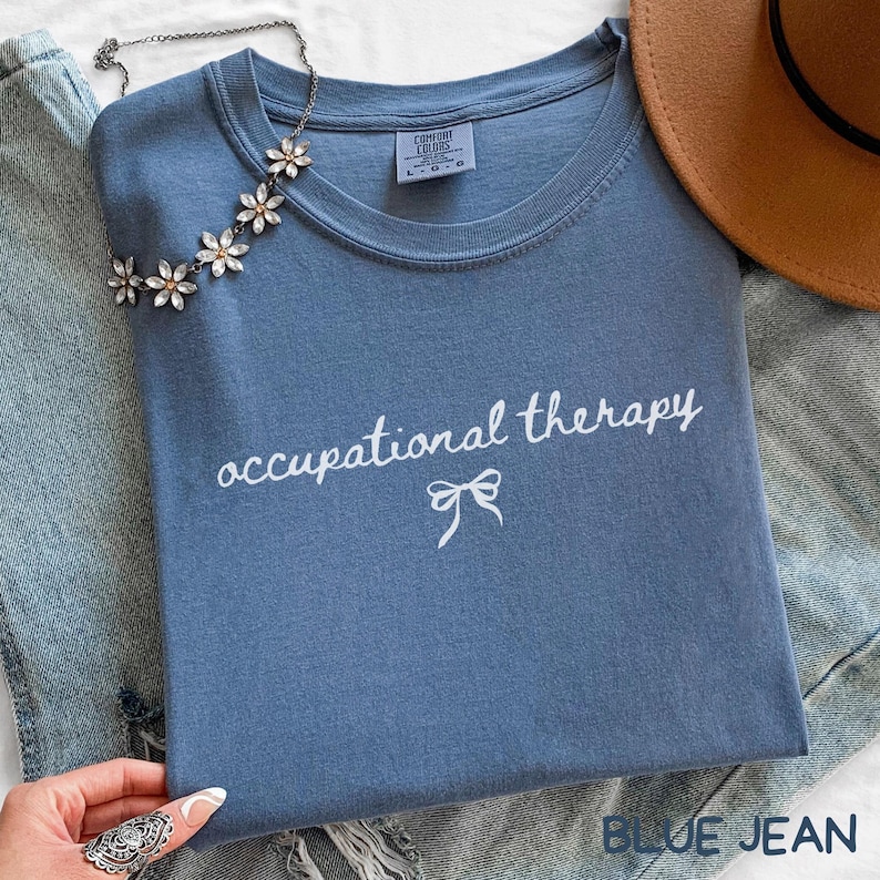 May include: A blue Comfort Colors t-shirt with the words "occupational therapy" in white script and a bow graphic. A floral necklace and a brown hat are also visible. The shirt is folded on top of a pair of blue jeans.