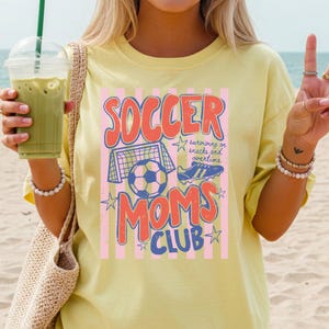 Puede incluir: Camiseta amarilla clara con un gr&aacute;fico que dice "SOCCER MOMS CLUB" en rojo y azul. El dise&ntilde;o incluye un bal&oacute;n de f&uacute;tbol, una porter&iacute;a y botas de f&uacute;tbol. La camiseta tambi&eacute;n tiene el texto "surviving on snacks and overtime".