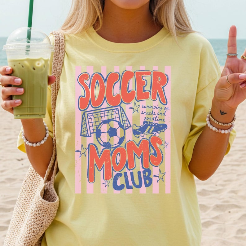 Puede incluir: Camiseta amarilla clara con un gr&aacute;fico que dice "SOCCER MOMS CLUB" en rojo y azul. El dise&ntilde;o incluye un bal&oacute;n de f&uacute;tbol, una porter&iacute;a y botas de f&uacute;tbol. La camiseta tambi&eacute;n tiene el texto "surviving on snacks and overtime".