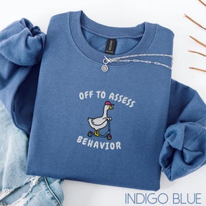 May include: Indigo blue sweatshirt with the phrase "OFF TO ASSESS BEHAVIOR" and a cartoon goose riding a scooter. A silver necklace with a pendant is draped across the sweatshirt. The sweatshirt is folded on top of a pair of blue jeans.