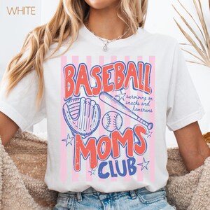 Op de afbeelding: Wit T-shirt met een honkbalthema. Het ontwerp toont het woord "BASEBALL" in het rood, een honkbalhandschoen, een honkbal en een knuppel. Daaronder staan de woorden "MOMS CLUB" in blauw. Het shirt bevat ook de tekst "surviving on snacks and homeruns."
