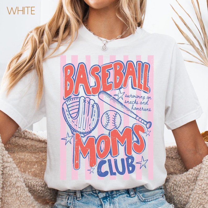 Op de afbeelding: Wit T-shirt met een honkbalthema. Het ontwerp toont het woord "BASEBALL" in het rood, een honkbalhandschoen, een honkbal en een knuppel. Daaronder staan de woorden "MOMS CLUB" in blauw. Het shirt bevat ook de tekst "surviving on snacks and homeruns."