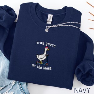 K&ouml;nnte beinhalten: Marineblaues Sweatshirt mit einem gestickten Design einer Gans mit M&uuml;tze, die einen Roller f&auml;hrt. Der Text "xray goose on the loose" befindet sich &uuml;ber und unter der Gans. Eine silberne Halskette ist dar&uuml;ber drapiert.