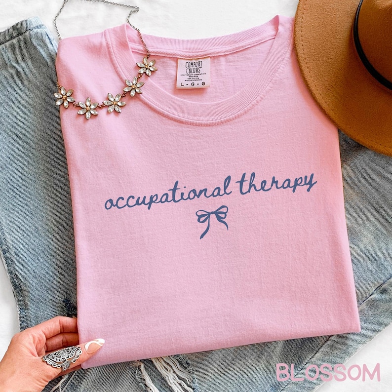 May include: A light pink t-shirt with the words "occupational therapy" in navy blue script and a bow graphic. The shirt has a crew neck and short sleeves. A silver necklace with flower-shaped accents is draped on the shirt. The word "BLOSSOM" is printed at the bottom.