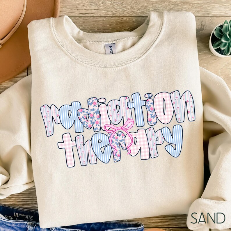 Może przedstawiać: Beżowa bluza z napisem "radiation therapy" w kwiatowy i kratkowy wz&oacute;r. Tekst jest niebieski i r&oacute;żowy z r&oacute;żową kokardą pośrodku. Bluza jest złożona.