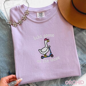 May include: A light purple t-shirt with the embroidered phrase "boba goose on the loose." The design features a cartoon goose wearing a pink cap and riding a blue scooter. A floral necklace and a brown hat are also visible.