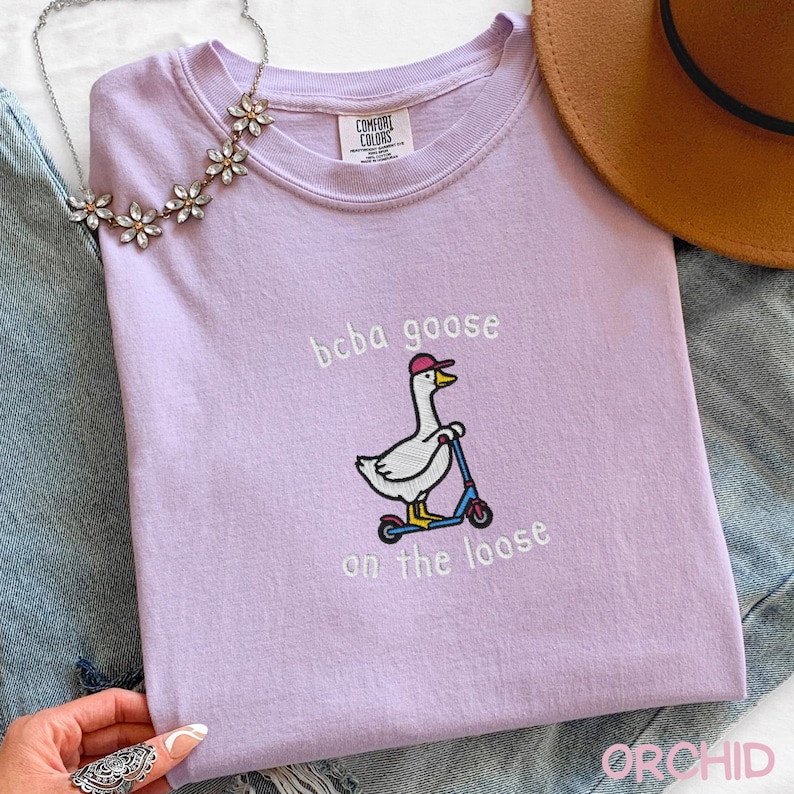 May include: A light purple t-shirt with the embroidered phrase "boba goose on the loose." The design features a cartoon goose wearing a pink cap and riding a blue scooter. A floral necklace and a brown hat are also visible.