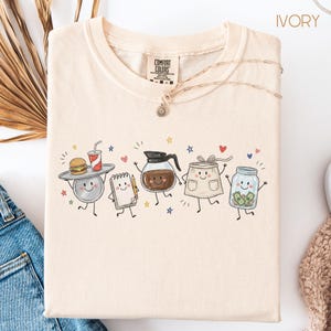May include: An ivory t-shirt featuring a graphic print of five anthropomorphic items: a tray with a burger and drink, a notepad with a pencil, a coffee pot, an apron, and a tip jar with money. The shirt has a Comfort Colors label at the neck.