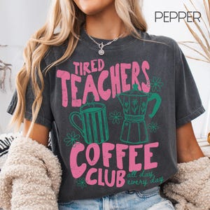 May include: Dark gray t-shirt with the text "Tired Teachers Coffee Club" in pink. The shirt features green illustrations of a coffee mug and a coffee maker. The words "all day every day" are also printed on the shirt.