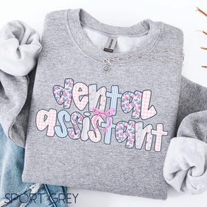 May include: A grey sweatshirt featuring the words "dental assistant" in a colourful, patterned design. The letters are decorated with floral, striped, and gingham patterns. A silver necklace with a small pendant is also visible.