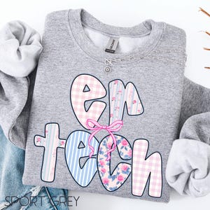 May include: A gray sweatshirt with the text "EM TECH" in a whimsical design. The letters are decorated with pink gingham, blue stripes, and floral patterns, tied together with a pink bow. A silver necklace with a small pendant is visible.