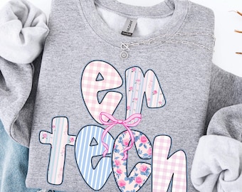 May include: A gray sweatshirt with the text "EM TECH" in a whimsical design. The letters are decorated with pink gingham, blue stripes, and floral patterns, tied together with a pink bow. A silver necklace with a small pendant is visible.