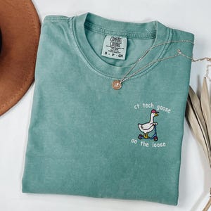 May include: A sage green short-sleeve t-shirt featuring an embroidered graphic of a white goose wearing a pink hat and riding a blue scooter. The text "ct tech goose on the loose" is embroidered in white around the goose graphic.