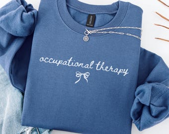 May include: Indigo blue sweatshirt with the words "occupational therapy" in white script, a bow graphic, and a silver necklace. The sweatshirt is folded on a white surface with a denim garment visible. The text "INDIGO BLUE" is at the bottom.