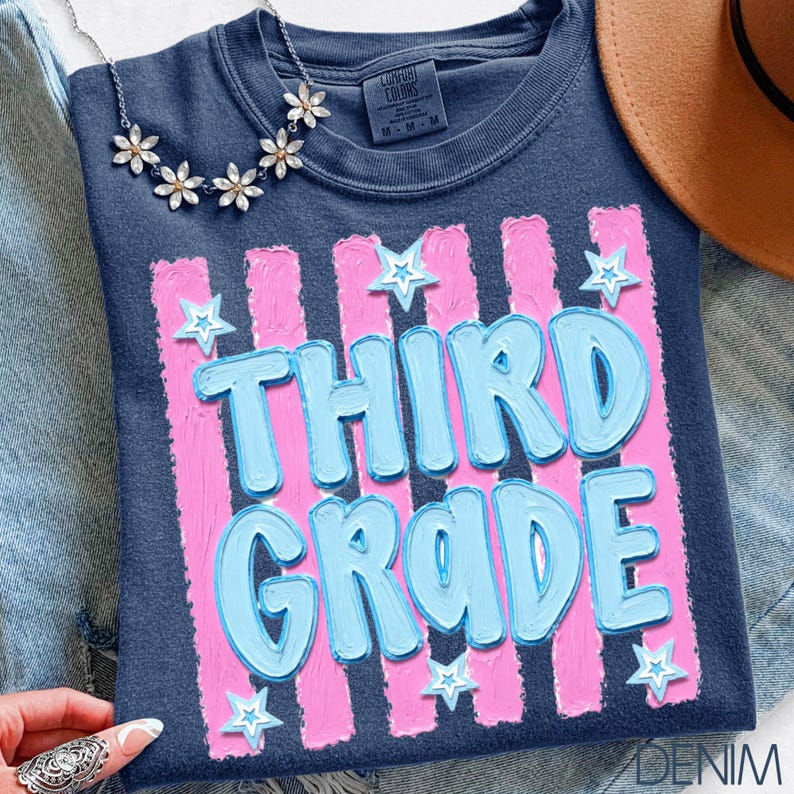 May include: Navy blue t-shirt with the words "THIRD GRADE" in light blue, outlined in a darker blue, set against pink vertical stripes. White star accents are scattered around the text. A silver necklace with flower accents is visible.