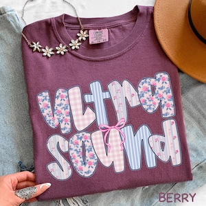 Op de afbeelding: Bessenkleurig T-shirt met de woorden "ULTRA SOUND" in een speels lettertype. De letters zijn versierd met bloemen-, ruit- en streepmotieven. Een roze strik accentueert het ontwerp. Een zilveren ketting met bloemhangers is ook zichtbaar.