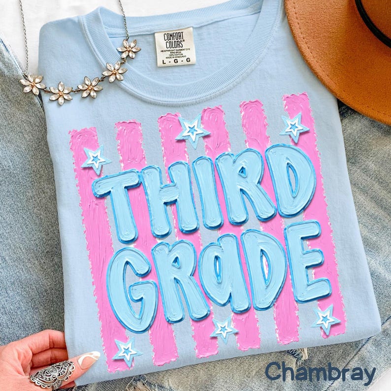 May include: Light blue t-shirt with the words "THIRD GRADE" in blue bubble letters, set against pink vertical stripes. White star accents are scattered around the text. The shirt is a Comfort Colors brand, size L-G.