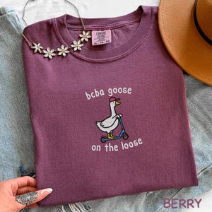 May include: Berry-colored t-shirt with the text "bcba goose on the loose" and a graphic of a goose riding a scooter. The shirt is a Comfort Colors brand, size XL-TG.