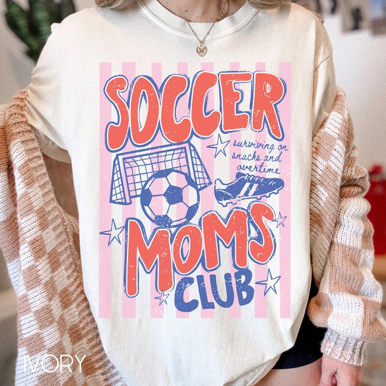 Puede incluir: Camiseta de color marfil con un dise&ntilde;o gr&aacute;fico. El dise&ntilde;o presenta las palabras "SOCCER MOMS CLUB" en rojo y azul, junto con ilustraciones de f&uacute;tbol. La camiseta tambi&eacute;n incluye el texto "surviving on snacks and overtime."