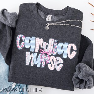 K&ouml;nnte beinhalten: Dunkelgraues Sweatshirt mit dem Schriftzug "Cardiac Nurse" in Gro&szlig;buchstaben. Die Buchstaben sind mit floralen, Vichy- und Streifenmustern verziert. Eine silberne Halskette mit Anh&auml;nger liegt &uuml;ber dem Sweatshirt.