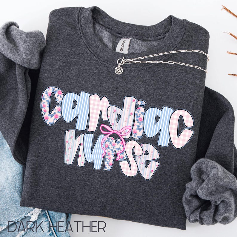 K&ouml;nnte beinhalten: Dunkelgraues Sweatshirt mit dem Schriftzug "Cardiac Nurse" in Gro&szlig;buchstaben. Die Buchstaben sind mit floralen, Vichy- und Streifenmustern verziert. Eine silberne Halskette mit Anh&auml;nger liegt &uuml;ber dem Sweatshirt.