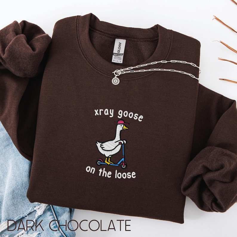 K&ouml;nnte beinhalten: Dunkelbraunes Sweatshirt mit dem gestickten Schriftzug "xray goose on the loose" und einer Cartoon-Gans auf einem Roller. Eine silberne Halskette mit Sonnenanh&auml;nger liegt auf dem Sweatshirt. Das Wort "Dark Chocolate" steht unten.