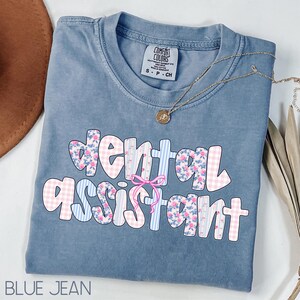 Pode incluir: Uma camiseta azul jeans Comfort Colors com as palavras "dental assistant" em um design floral e xadrez. Um colar de ouro com um pingente est&aacute; drapeado sobre a camisa. A camisa est&aacute; dobrada cuidadosamente.