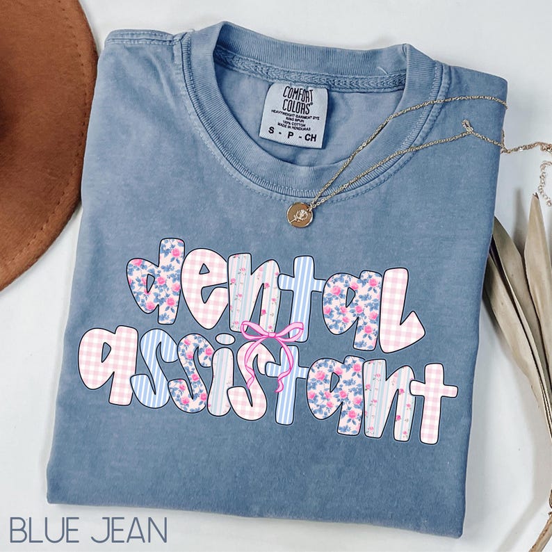 Pode incluir: Uma camiseta azul jeans Comfort Colors com as palavras "dental assistant" em um design floral e xadrez. Um colar de ouro com um pingente est&aacute; drapeado sobre a camisa. A camisa est&aacute; dobrada cuidadosamente.
