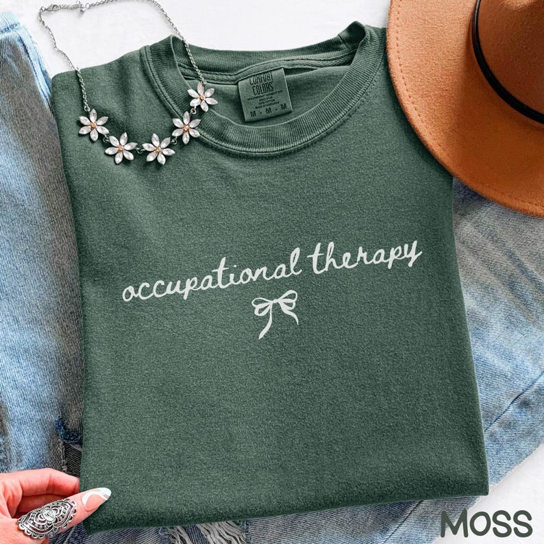 May include: A moss green sweatshirt with the words "occupational therapy" in white script and a small bow graphic. A silver flower necklace and a brown hat are also visible.