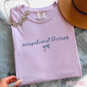 May include: A lilac-colored t-shirt with the words "occupational therapy" in a dark blue script, with a bow graphic underneath. The shirt is made of a soft material and is part of a clothing collection.