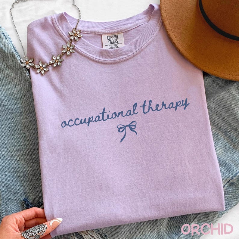 May include: A lilac-colored t-shirt with the words "occupational therapy" in a dark blue script, with a bow graphic underneath. The shirt is made of a soft material and is part of a clothing collection.
