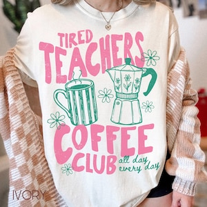 May include: Ivory-colored t-shirt with a pink and green graphic. The graphic includes the words "Tired Teachers Coffee Club" with a coffee mug, a coffee pot, and floral accents. The phrase "all day every day" is also included.