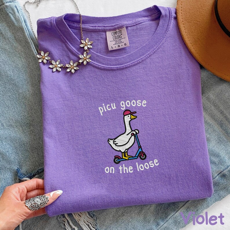 K&ouml;nnte beinhalten: Violettes Comfort Colors T-Shirt mit dem Text "picu goose on the loose" und einer gestickten Gans auf einem Roller. Das Shirt hat einen Rundhalsausschnitt und kurze &Auml;rmel.