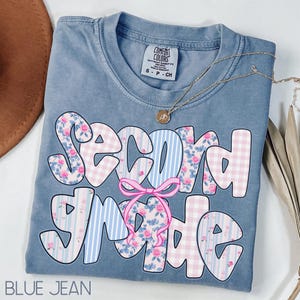 Peut inclure: T-shirt bleu jean Comfort Colors avec l'inscription "Second Grade" dans une police fantaisiste. Les lettres présentent des motifs floraux, vichy et rayés, avec un nœud rose. Un collier doré est drapé sur le t-shirt.