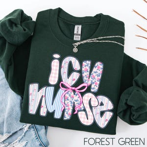 May include: Forest green sweatshirt with the text "ICU Nurse" in a decorative font. The letters are patterned with pink gingham, blue stripes, and floral designs. A pink bow is between the "C" and "N". A silver necklace is draped across the sweatshirt.