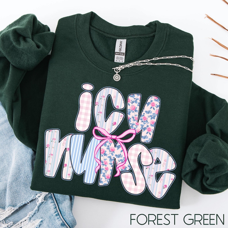May include: Forest green sweatshirt with the text "ICU Nurse" in a decorative font. The letters are patterned with pink gingham, blue stripes, and floral designs. A pink bow is between the "C" and "N". A silver necklace is draped across the sweatshirt.