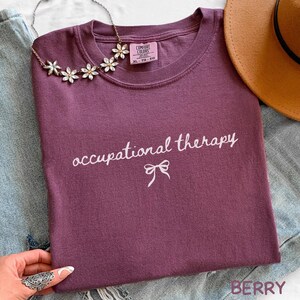 May include: A berry-colored t-shirt with the words "occupational therapy" in white script and a bow graphic. A floral necklace and a brown hat are also visible. The shirt is folded on a denim surface.