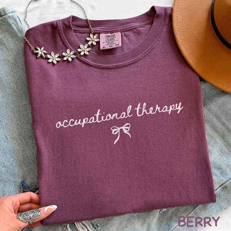 May include: A berry-colored t-shirt with the words "occupational therapy" in white script and a bow graphic. A floral necklace and a brown hat are also visible. The shirt is folded on a denim surface.