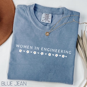 Peut inclure: Un t-shirt bleu jean avec le texte "WOMEN IN ENGINEERING" et une rangée de marguerites blanches. Le t-shirt a un aspect vieilli et est fabriqué par Comfort Colors.