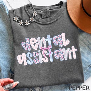 Pode incluir: T-shirt cinza escuro com as palavras "dental assistant" em um design colorido e estampado. O texto apresenta padr&otilde;es florais, xadrez e listrados em rosa, azul e branco. Um la&ccedil;o rosa est&aacute; centralizado acima da palavra "assistant".
