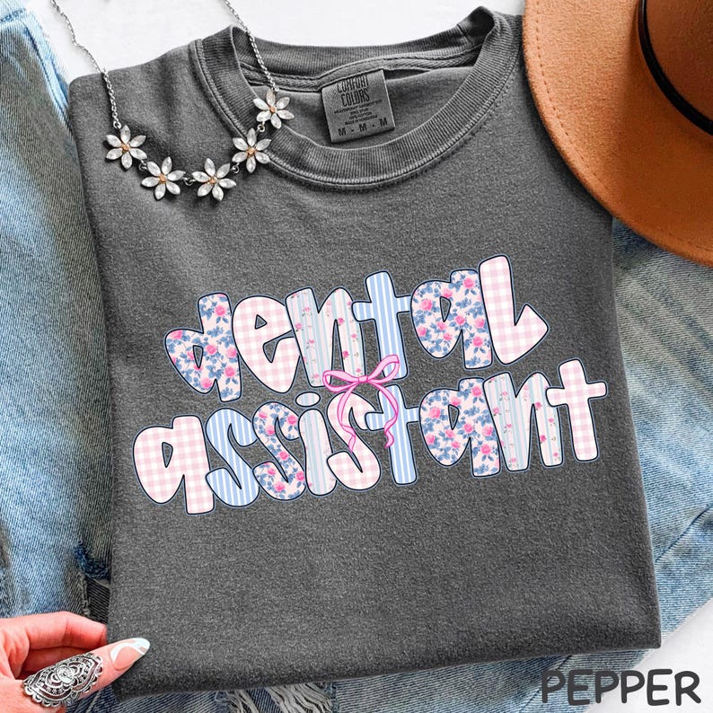 Pode incluir: T-shirt cinza escuro com as palavras "dental assistant" em um design colorido e estampado. O texto apresenta padr&otilde;es florais, xadrez e listrados em rosa, azul e branco. Um la&ccedil;o rosa est&aacute; centralizado acima da palavra "assistant".
