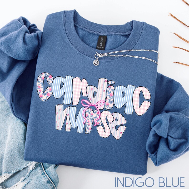 K&ouml;nnte beinhalten: Indigoblaues Sweatshirt mit Rundhalsausschnitt und dem Schriftzug "Cardiac Nurse" in einer verspielten Schriftart. Die Buchstaben sind mit floralen und gestreiften Mustern in Rosa und Blau verziert. Eine silberne Halskette mit einem kleinen Anh&auml;nger ist dar&uuml;ber drapiert.