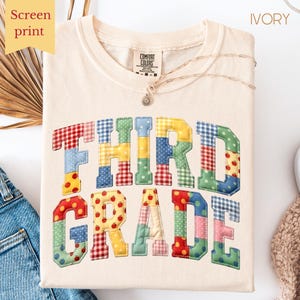 Peut inclure: T-shirt couleur ivoire avec l'inscription "THIRD GRADE" dans un motif patchwork. Les lettres sont composées de divers tissus à motifs, dont du vichy, des pois et des imprimés de pommes. Un collier délicat repose sur le t-shirt.
