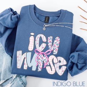 May include: Indigo blue crewneck sweatshirt with the text "ICU Nurse" in a whimsical font. The letters are decorated with floral and gingham patterns, and a pink bow. A silver necklace with a small pendant is draped across the sweatshirt.