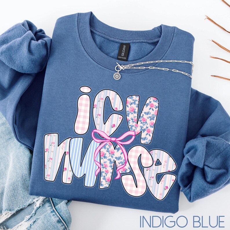 May include: Indigo blue crewneck sweatshirt with the text "ICU Nurse" in a whimsical font. The letters are decorated with floral and gingham patterns, and a pink bow. A silver necklace with a small pendant is draped across the sweatshirt.