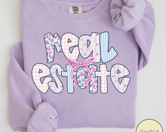 May include: A light purple sweatshirt with the words "REAL ESTATE" in a bubble font. The letters are decorated with floral, gingham, and striped patterns. A pink bow is centered between the words. A scrunchie rests on the shoulder.