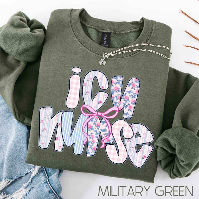 May include: Military green sweatshirt with the text "ICU Nurse" in a decorative font. The letters are patterned with pink gingham, floral, and blue striped designs. A pink bow is centered between the letters. A silver necklace with a sunburst pendant is on the sweatshirt.
