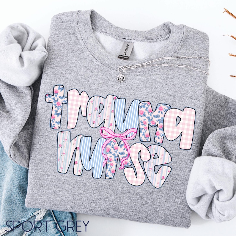 Puede incluir: Una sudadera gris con la frase "Trauma Nurse" en fuente de burbujas. Las letras est&aacute;n decoradas con estampados florales, cuadros y rayas en rosa, azul y blanco. Un lazo rosa est&aacute; entre las palabras.