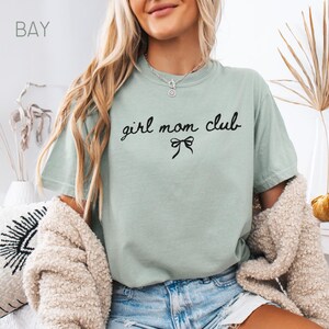 Peut inclure: Un t-shirt vert clair avec le texte "girl mom club" en &eacute;criture noire avec un n&oelig;ud noir en dessous.